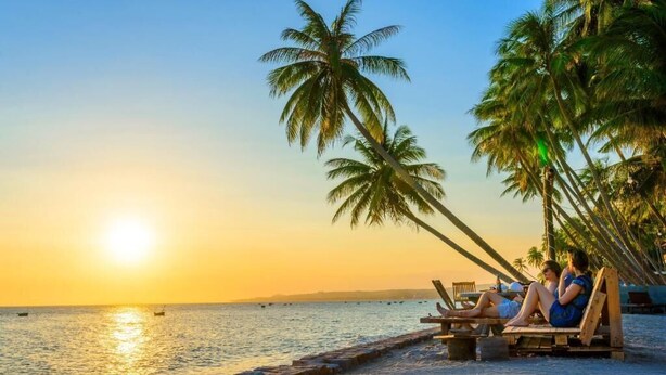 Een strandscène met palmbomen, een zonsondergang en twee personen die ontspannen in houten stoelen.