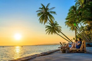 A beach scene with palm trees, a sunset, and two people relaxing on wooden chairs. - iHome Mui Ne - Hostel (Mui Ne)