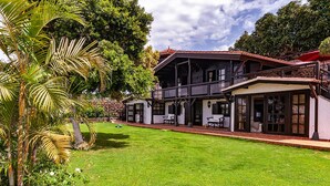 A two-story house with a balcony, a well-maintained lawn, and a garden with palm trees.