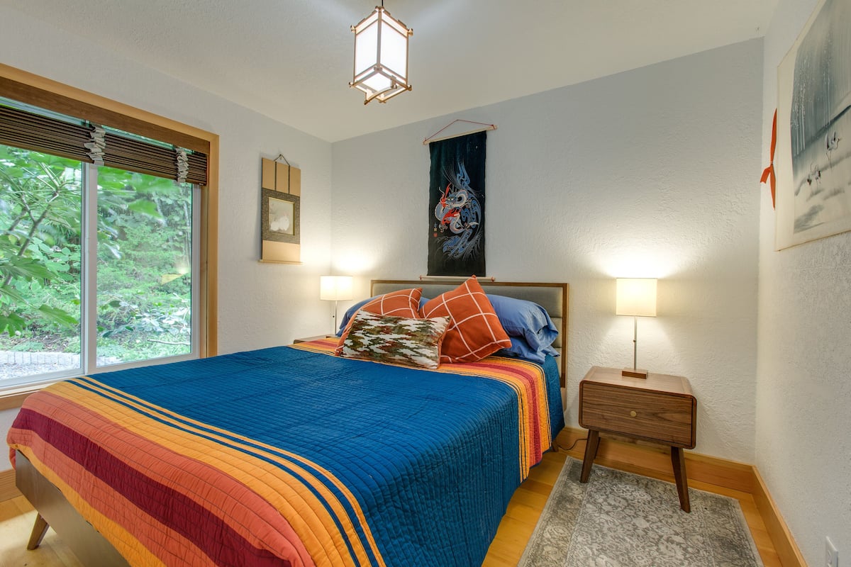 A bedroom with a large bed, two bedside tables, a nightstand, a window with blinds, a hanging tapestry, and a wall-mounted lamp.