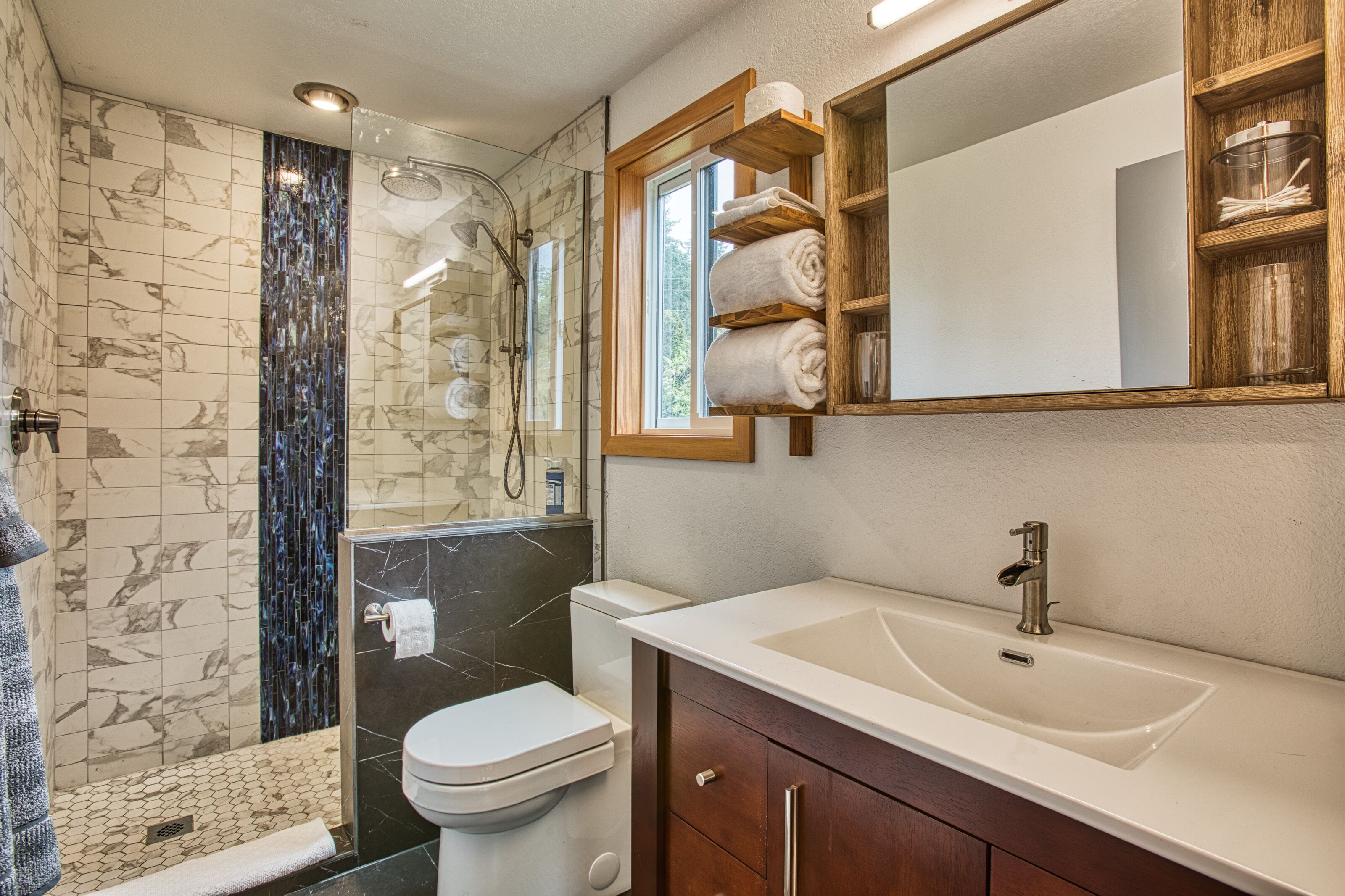 Un baño moderno con ducha de vidrio, inodoro blanco, un lavabo grande y repisas de madera sobre el espejo.
