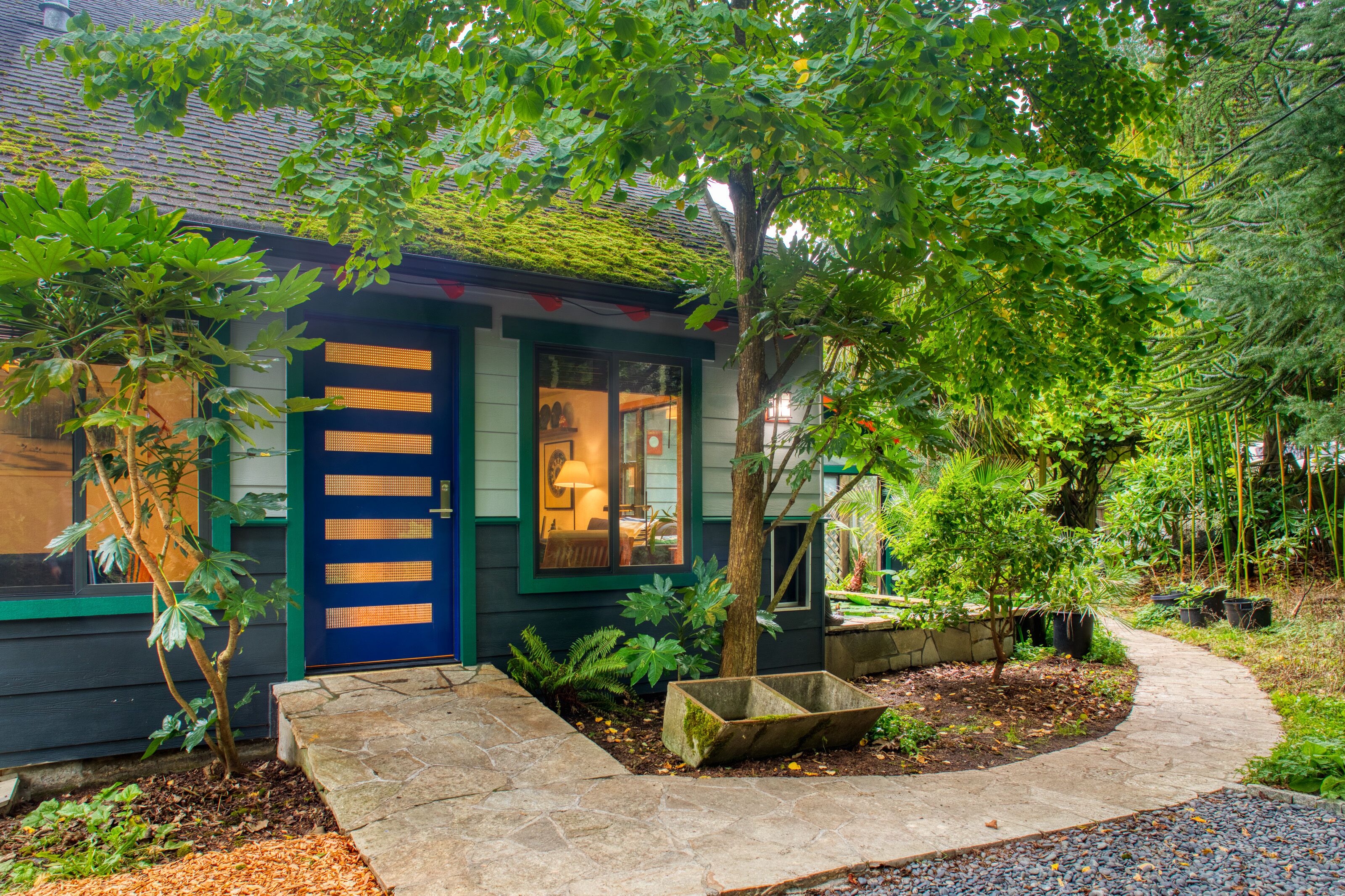 A house with a blue door, a visible interior with a lamp, and a garden with various plants and a stone pathway. - Sehome Garden Inn (Bellingham)