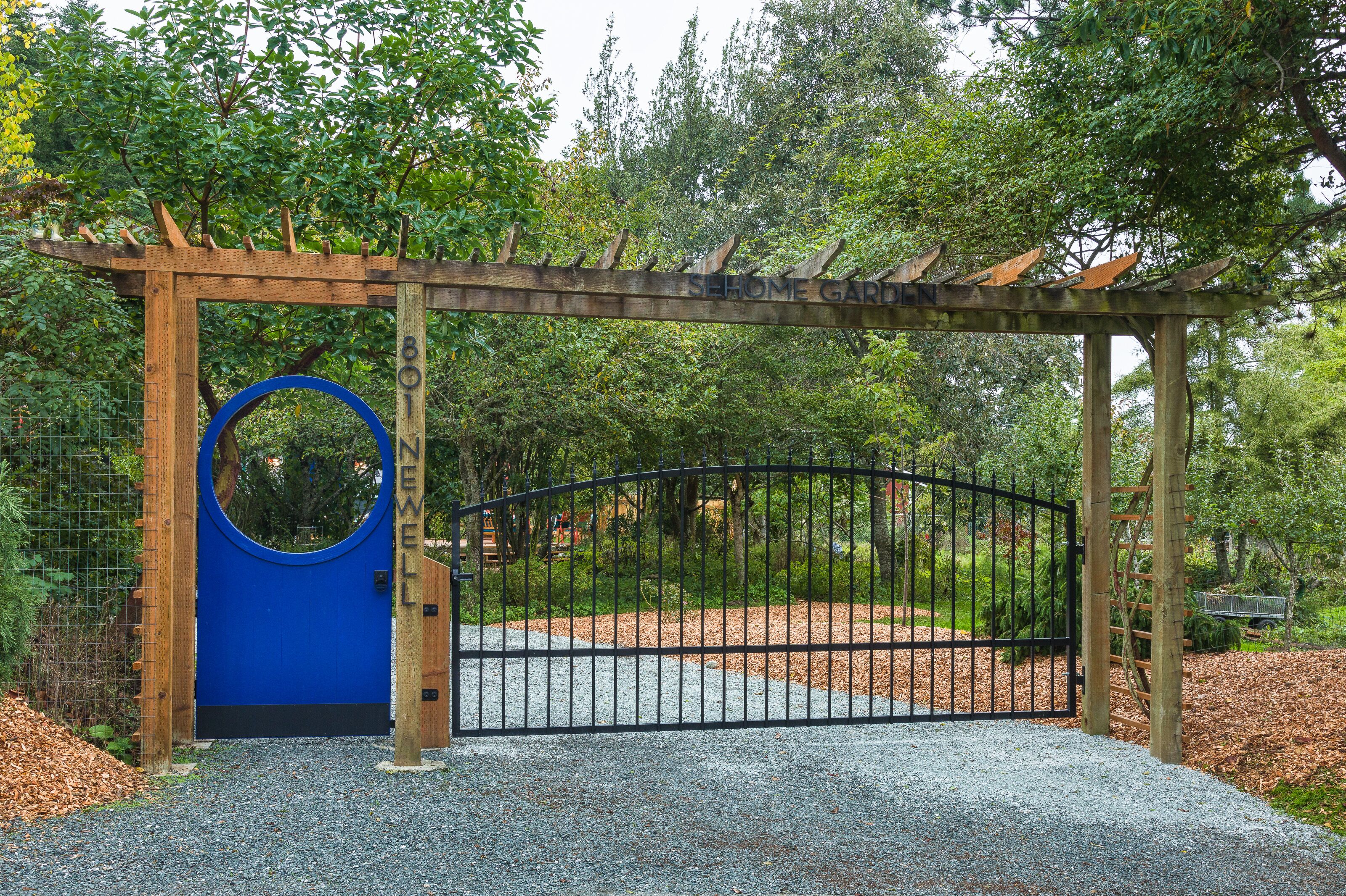 A wooden pergola entrance with a blue circular feature and a black gate, surrounded by greenery.