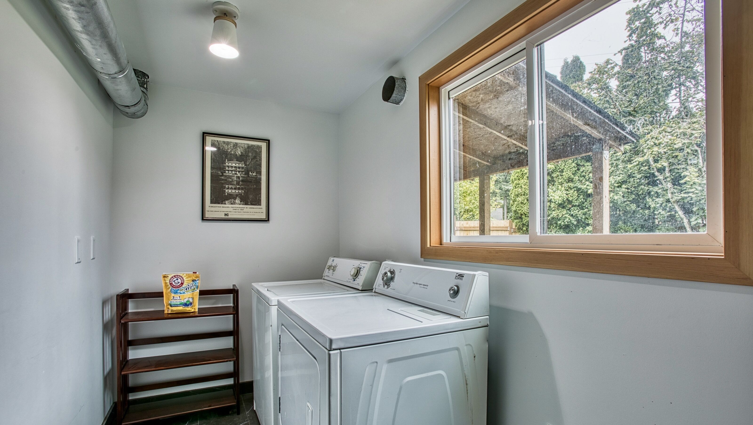 A laundry room with two stacked washers and dryers, a wooden shelf with a bag of detergent, a framed historical photo on the wall, and a window with a view of trees.