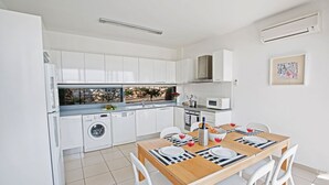 Une cuisine moderne avec des meubles blancs, un lave-linge encastré et un coin repas comprenant une table en bois et des chaises blanches.