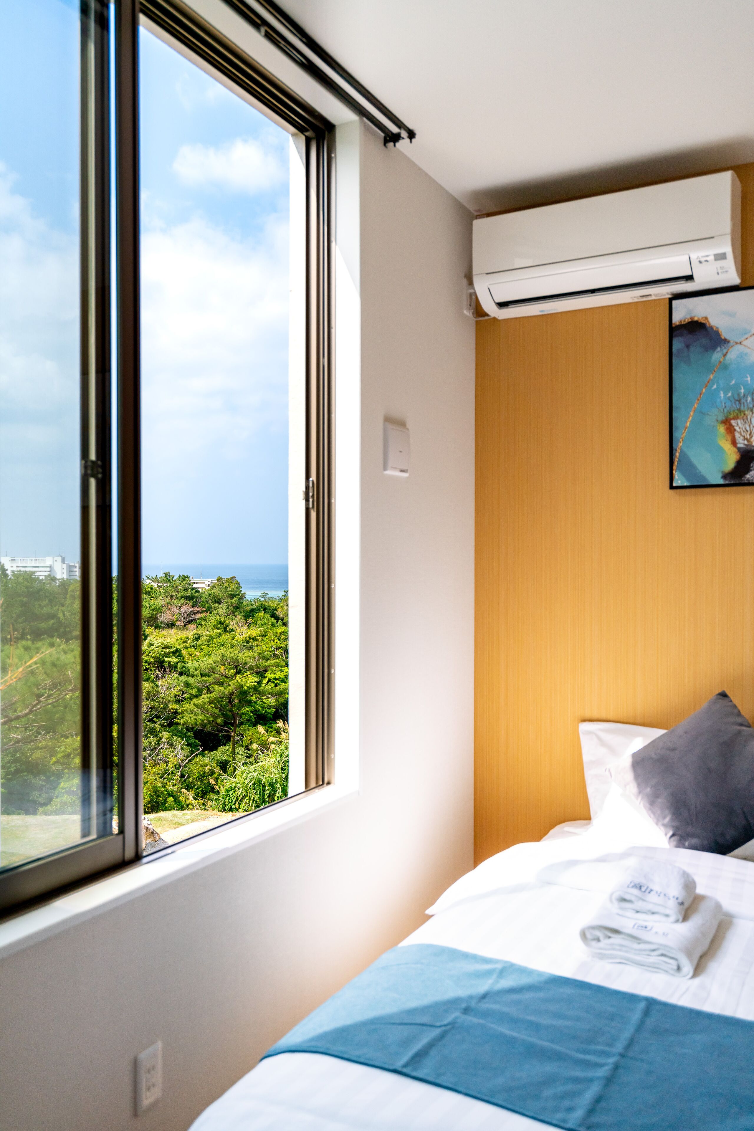 Ein Hotelzimmer mit großem Fenster, einem Bett mit weißer und blauer Bettwäsche, einem grauen Kopfkissen und einer Klimaanlage an der Wand.