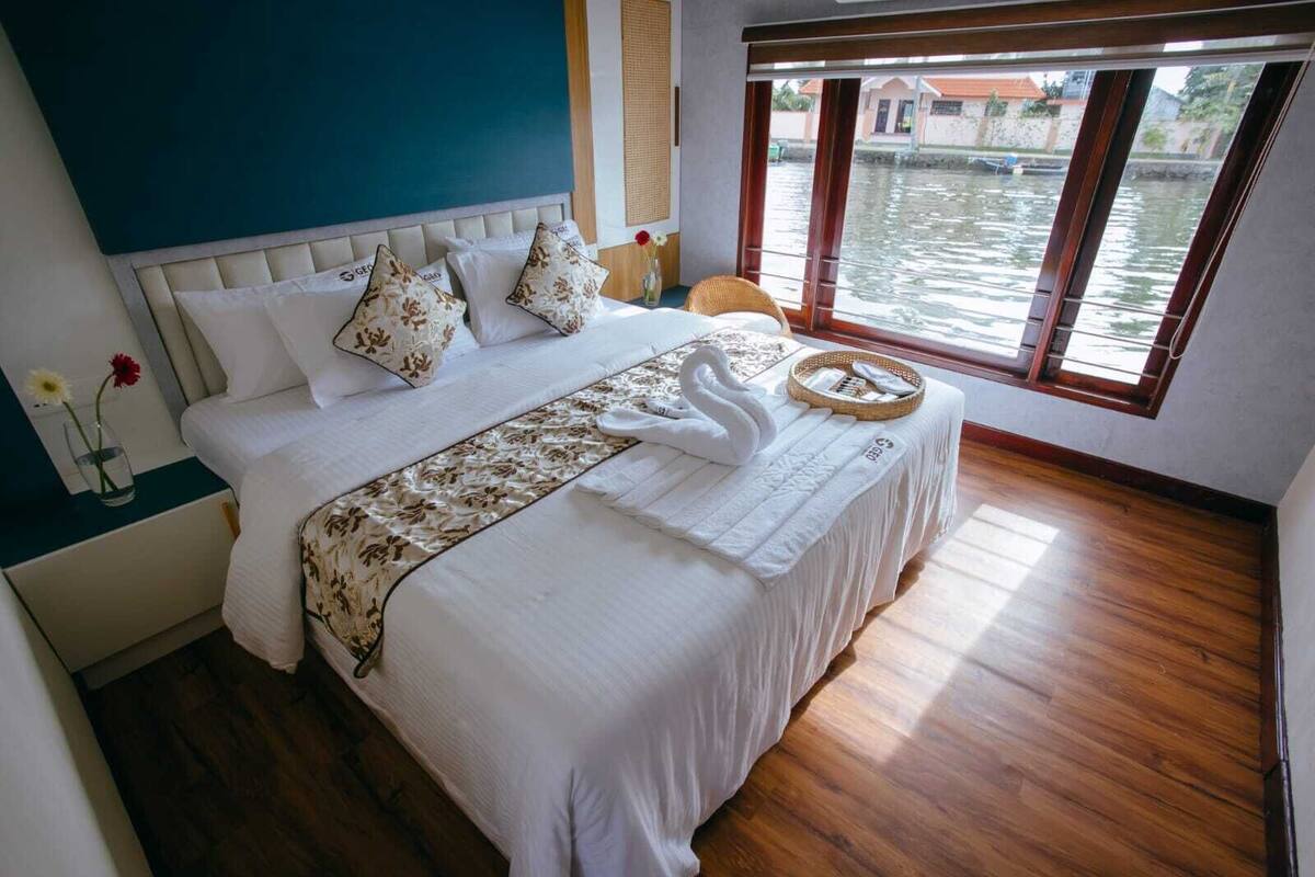 A bedroom with a bed, pillows, a bedside table with a vase, and a window with a view of water.