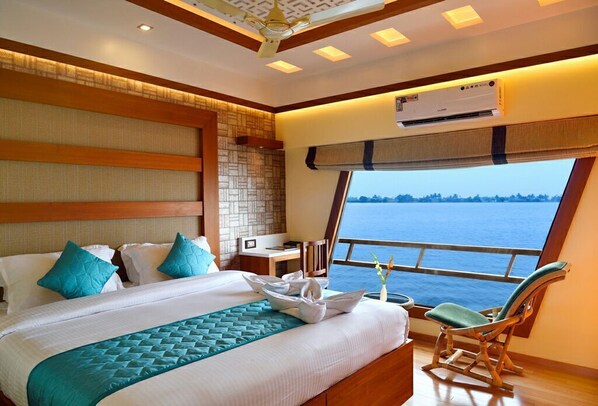 A hotel room with a large bed, a wooden headboard, a window with a view of the sea, and a ceiling fan.