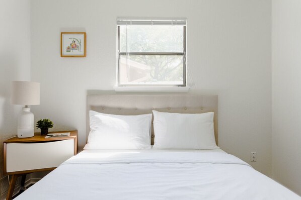 Une chambre d’hôtel avec un lit, du linge de lit blanc, une table de chevet avec une lampe, une petite plante et une fenêtre avec des stores.