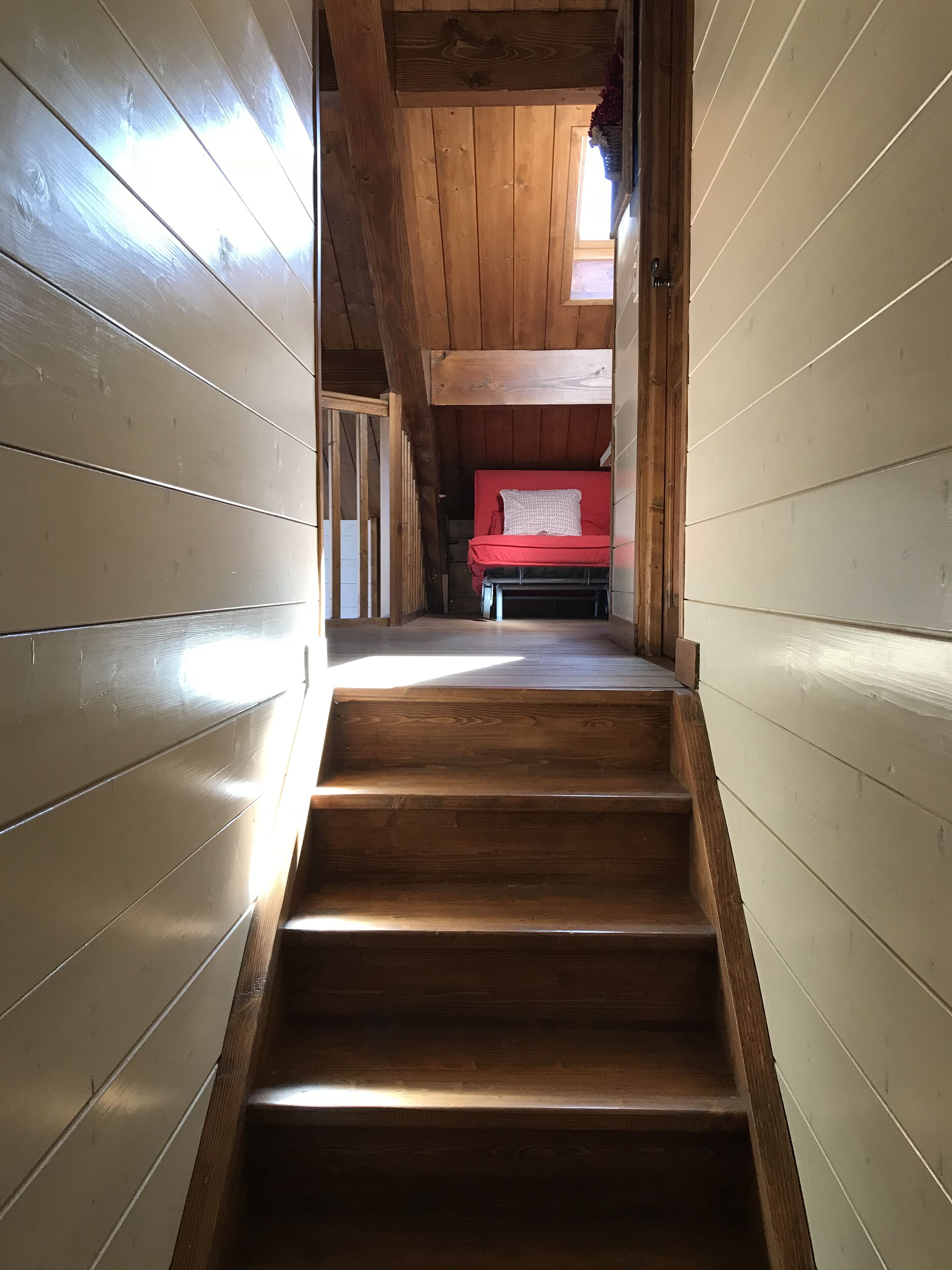 Eine Holztreppe führt zu einem roten Bett in einem Blockhausszimmer mit Holzwänden und einer geneigten Decke.