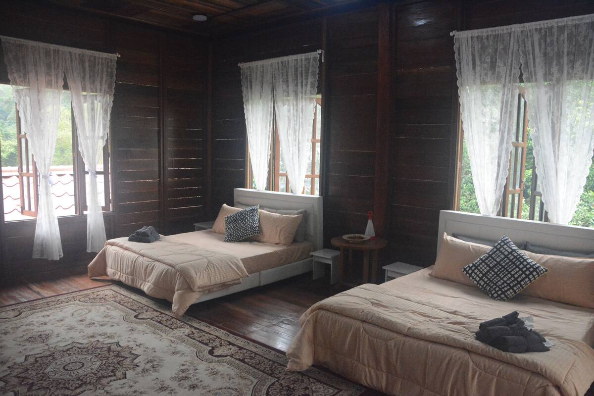 Una habitación con dos camas, cada una con una almohada y una manta. Hay una mesita con una lámpara y una ventana con cortinas.
