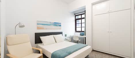 Un dormitorio con cama, una silla, un escritorio, un armario y un cuadro de una playa.