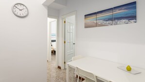 Un couloir avec une horloge murale blanche, un coin repas avec une table et des chaises blanches, et une porte donnant sur une autre pièce.