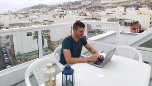 Un homme travaille sur un ordinateur portable sur un balcon, avec une vue sur le paysage urbain.