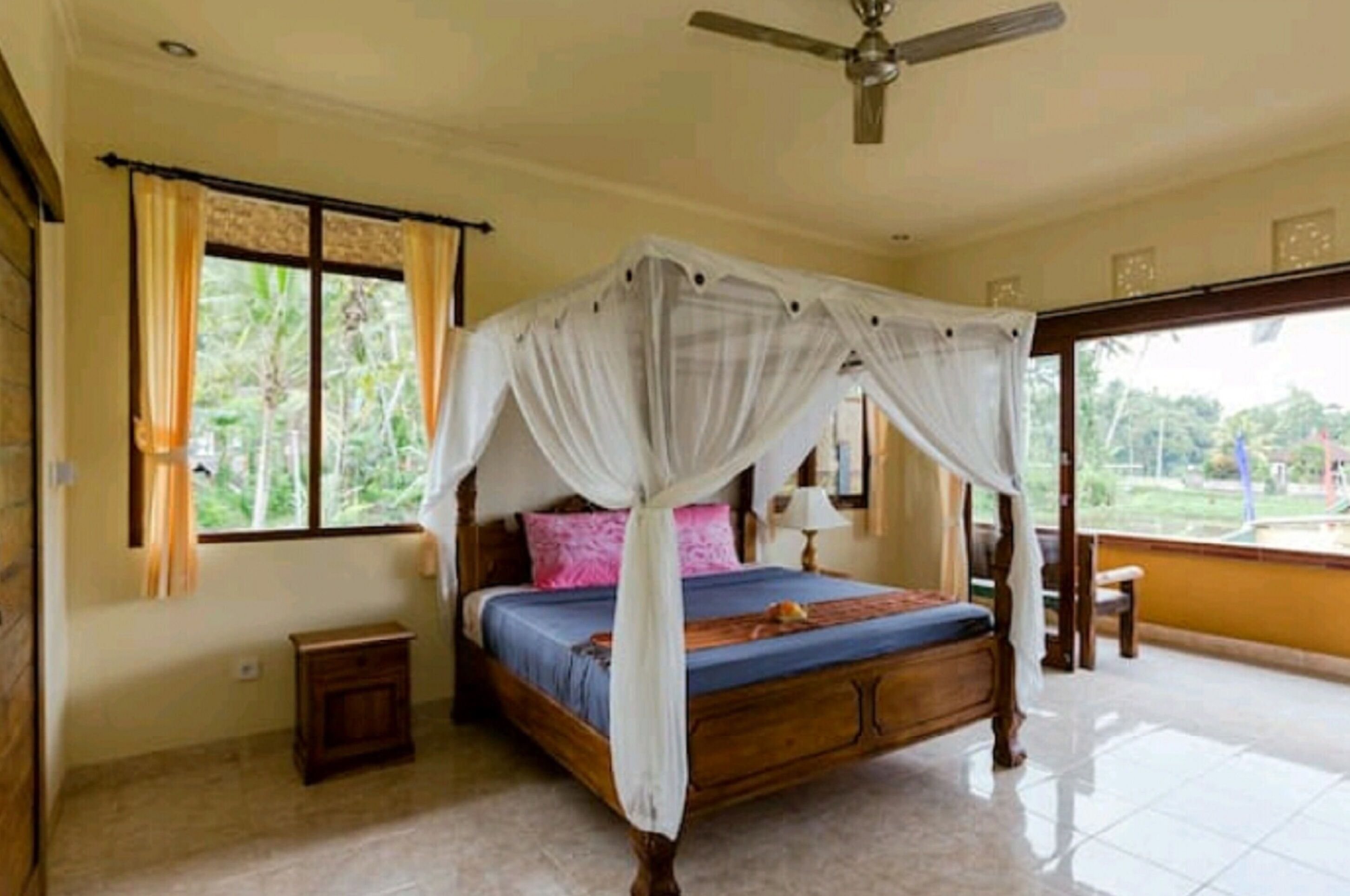 A bedroom with a four-poster bed, a ceiling fan, and a view of greenery outside.