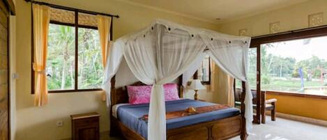 A bedroom with a four-poster bed, a ceiling fan, and a view of greenery outside.