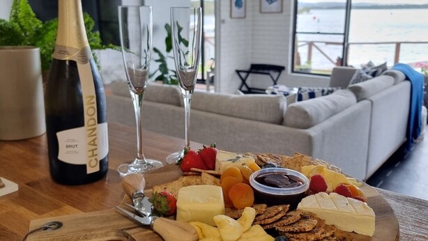 Une table en bois, sur laquelle on a disposé une planche de charcuterie, une bouteille de vin brut et deux verres à vin.