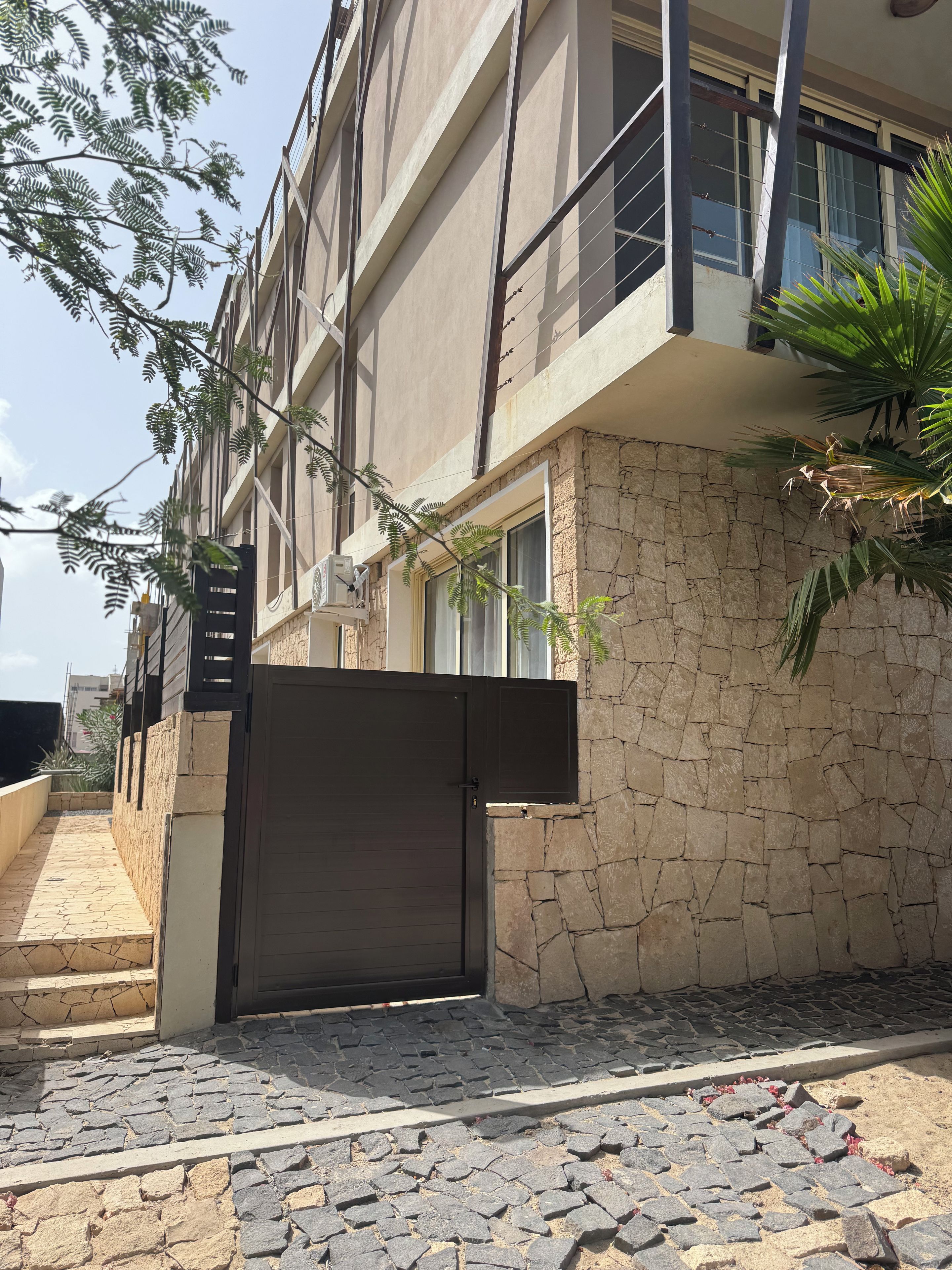 Un edificio de varios pisos con fachada de piedra, entrada protegida por una reja y un camino de acceso pavimentado.