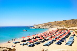 A beach with rows of sun loungers and umbrellas, a rocky hillside, and a clear blue sea. - Mykonian Studios (Mykonos)