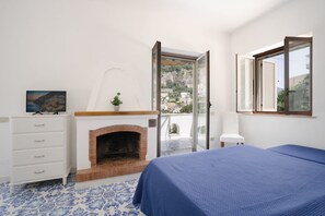 A bedroom with a fireplace, a bed, a window with shutters, and a view of a town. - Villa Carrino (Positano)