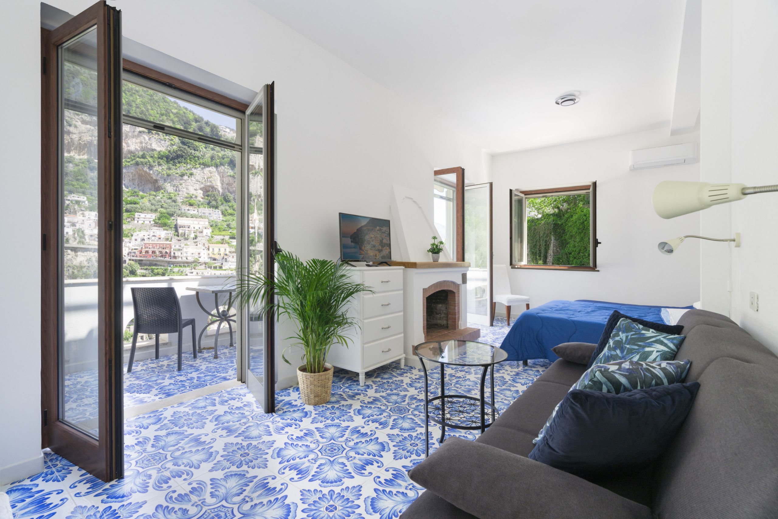 A room with a blue patterned floor, a grey sofa, a small table, a dresser, a mirror, a bed with a blue bedspread, and a balcony with a view of buildings and greenery.
