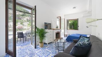 A room with a blue patterned floor, a grey sofa, a small table, a dresser, a mirror, a bed with a blue bedspread, and a balcony with a view of buildings and greenery.