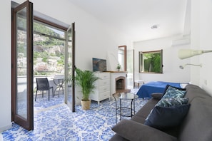 Een kamer met een blauw patroon vloer, een grijze bank, een kleine tafel, een dressoir, een spiegel, een bed met een blauw dekbed en een balkon met uitzicht op gebouwen en groen.