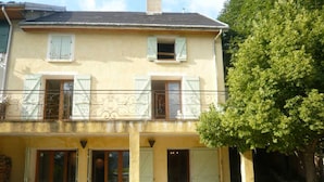 A two-story building with a balcony, outdoor seating area with white plastic chairs, and a tree to the right.