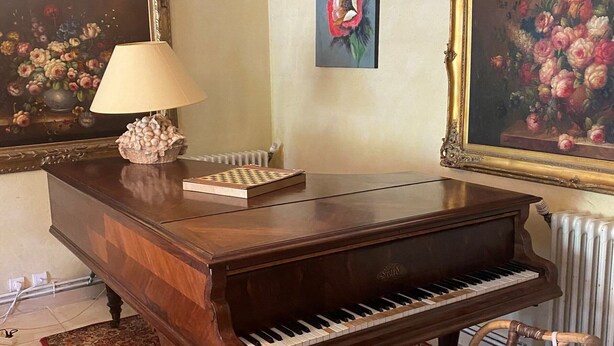 A grand piano, a chessboard, a lamp, and a chair in a room with a floral painting on the wall.