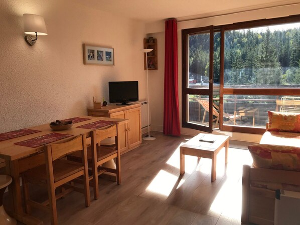 Ein gemütliches Zimmer mit einem Holzesstisch, Stühlen, einem Fernseher und einem großen Fenster mit Blick auf einen Wald und See.
