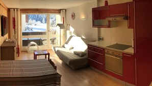 Ein kompakter Wohnraum mit einer Küchenzeile, einem Sofa, einem Esstisch und Blick auf eine verschneite Landschaft durch eine Glasdoor.
