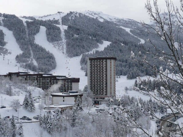 A snow-covered mountain resort with a tall building and several smaller structures, surrounded by trees and a ski slope.
