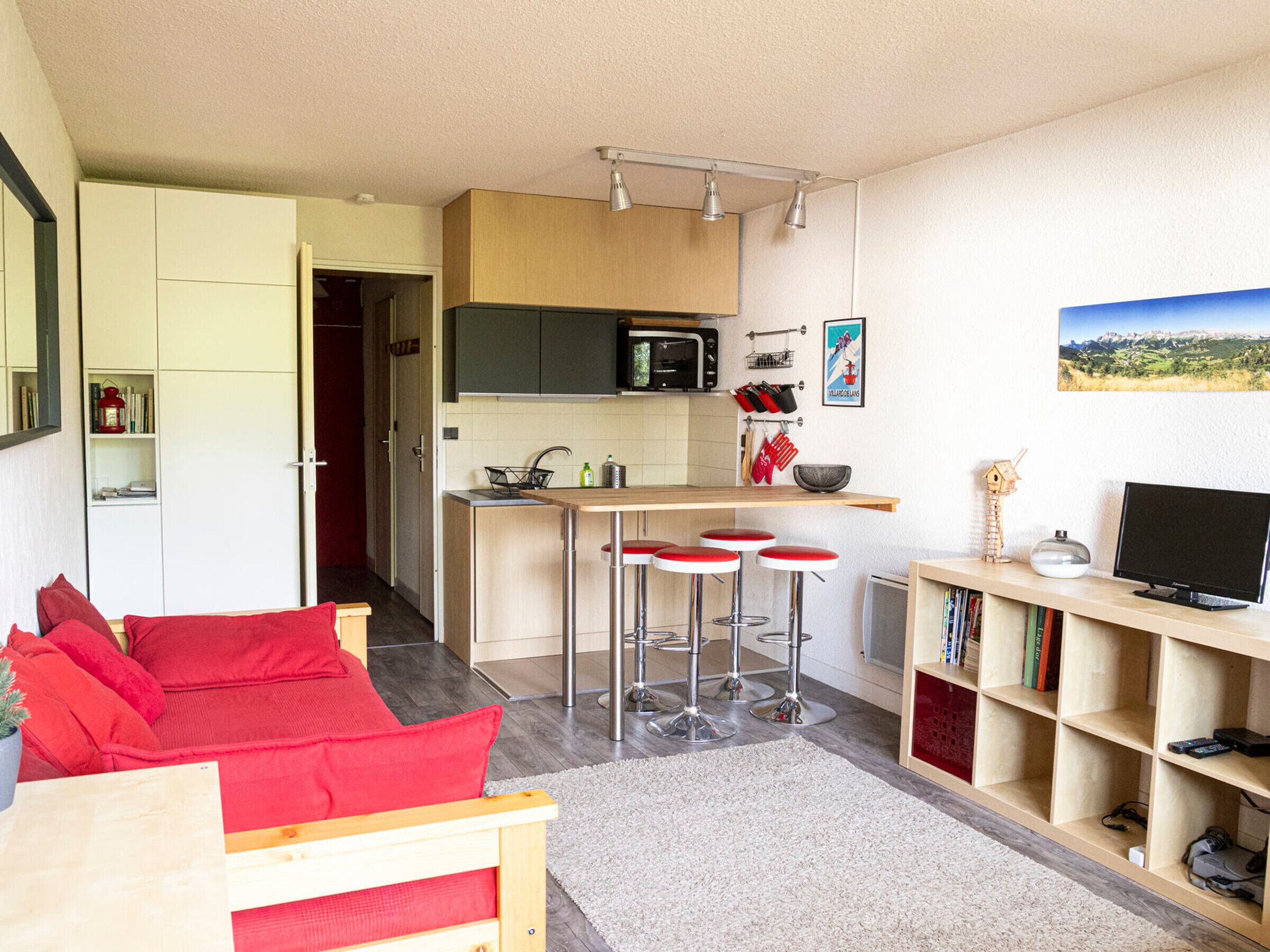 Een knus woongedeelte met een kitchenette, een eethoek met rode barstoelen, een boekenkast en een televisie.