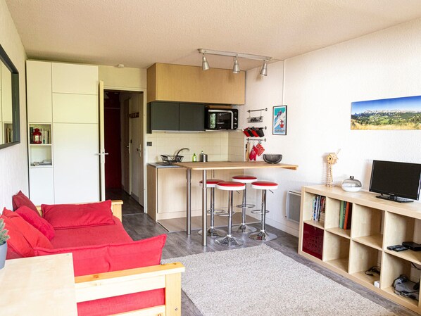 Un espace de vie compact comprenant un coin cuisine, un espace repas avec des tabourets de bar rouges, une bibliothèque et un téléviseur.