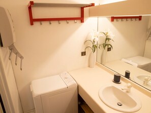 Une salle de bain avec un lavabo blanc, un miroir, un porte-serviettes et un petit vase blanc contenant des fleurs.