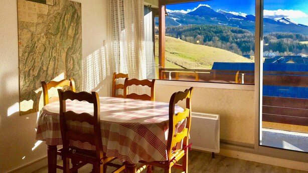 A dining area with a table and chairs, a map on the wall, and a large window with a mountain view. - Studio cabin with balcony, mountain view near village center (Villard-de-Lans)