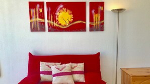A red sofa with patterned pillows, a wooden side table, and three abstract paintings on the wall. - Studio cabin with balcony, mountain view near village center (Villard-de-Lans)
