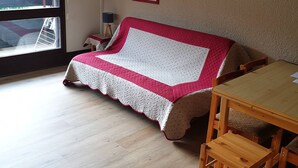 A cozy room with a wooden floor, a red and white quilted sofa, a wooden table, and a window with a view of trees. - 32m² Studio at Ski Slopes Foot in Corrençon (Corrençon-en-Vercors)