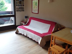 A cozy room with a wooden floor, a red and white quilted sofa, a wooden table, and a window with a view of trees.