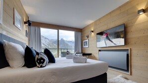 Une chambre moderne avec un grand lit, un téléviseur fixé au mur et une vue sur les montagnes grâce à de grandes fenêtres.