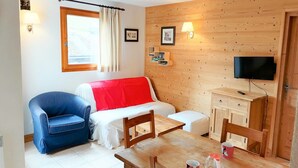 A cozy room with a bed, a blue chair, a wooden table, a TV, and a window with curtains. - 3-room apartment in Morillon 1100, 6 people, pets allowed, wifi (Morillon 1100)