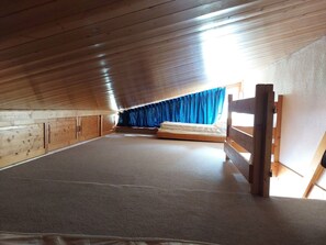 Quarto de sótão com pé-direito alto, uma cama beliche de madeira, uma janela com cortinas azuis e teto inclinado.