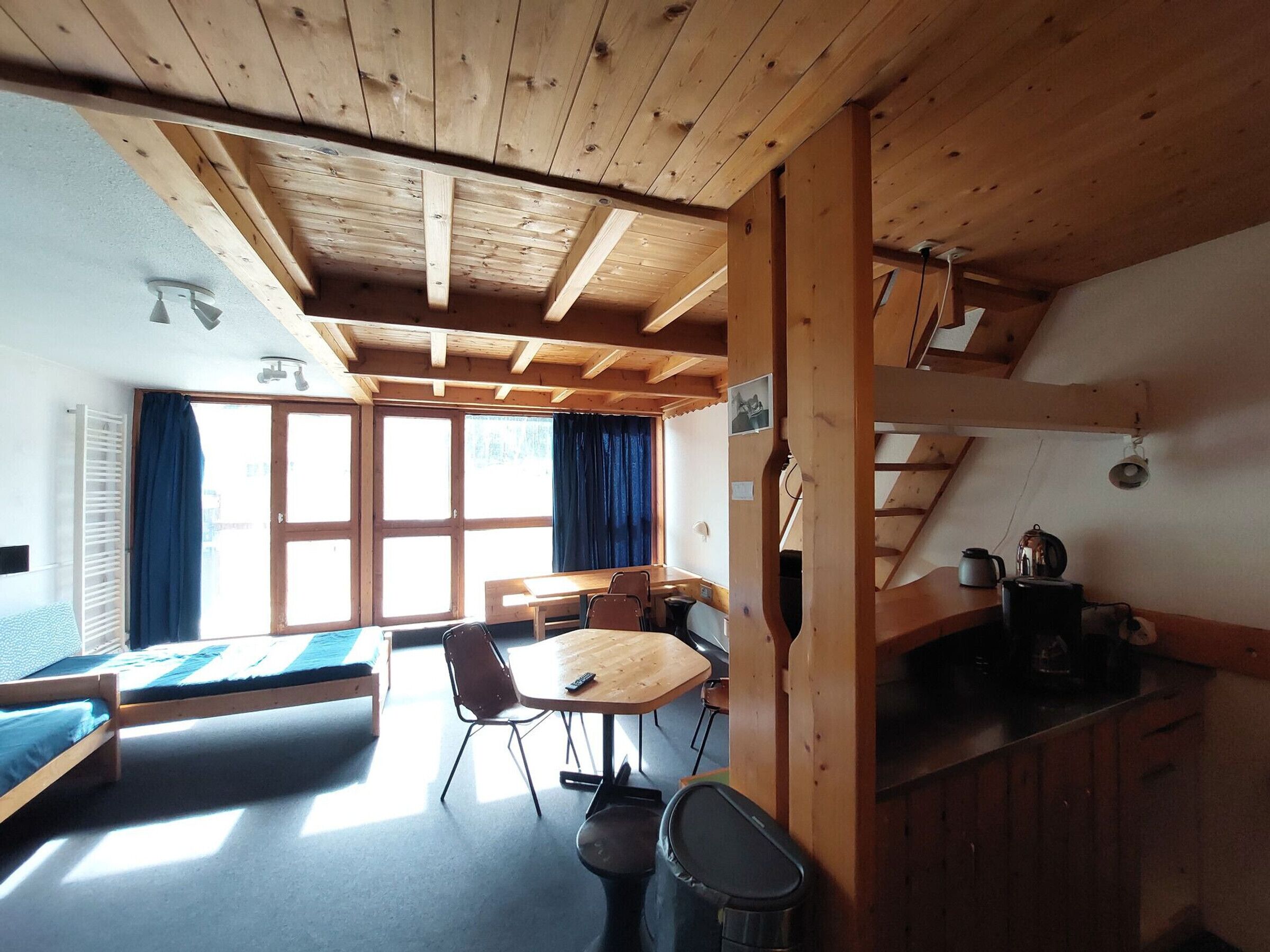 A wooden interior with a bed, a table, chairs, a bench, and a kitchen area.
