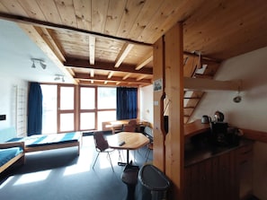 A wooden cabin interior with a bed, a dining table, chairs, a kitchen area, and a staircase.