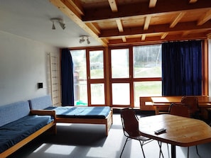 A room with a bed, a sofa, a table, and chairs. There are large windows with curtains.