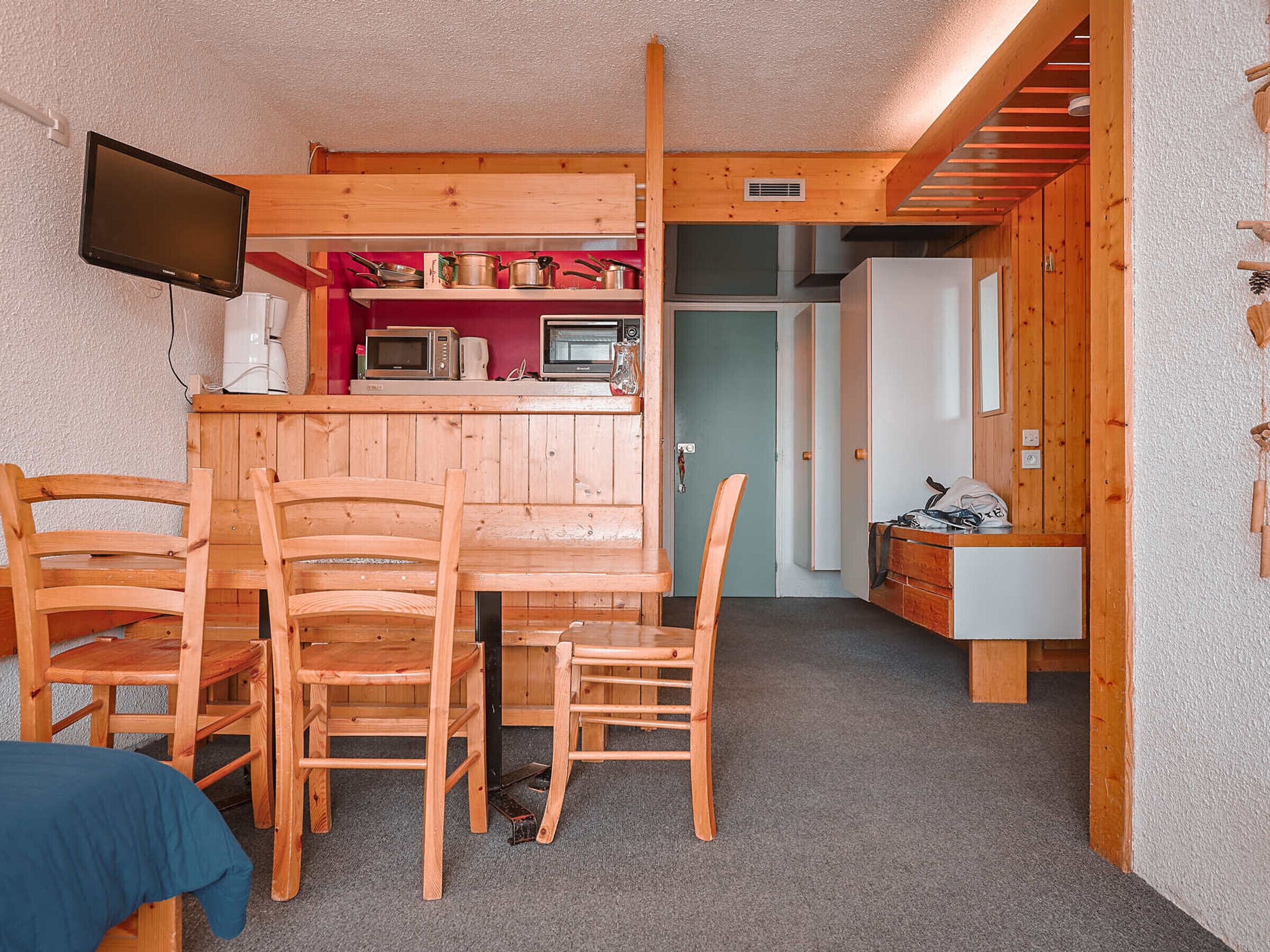 Une cuisine compacte avec des meubles en bois, un four à micro-ondes et un réfrigérateur. On y trouve une table à manger avec des chaises et un téléviseur fixé au mur.