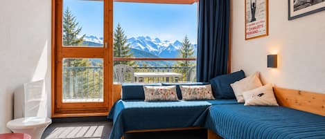 A bedroom with a bed, a small table, a red chair, a white toilet, and a balcony with a mountain view.
