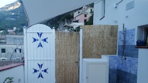 Ein Balkon mit einer weißen Tür, verziert mit blauen Sterndrucken, einem Bambuszaun, einem kleinen weißen Schrank und einem Duschbereich mit blauen Fliesen.
