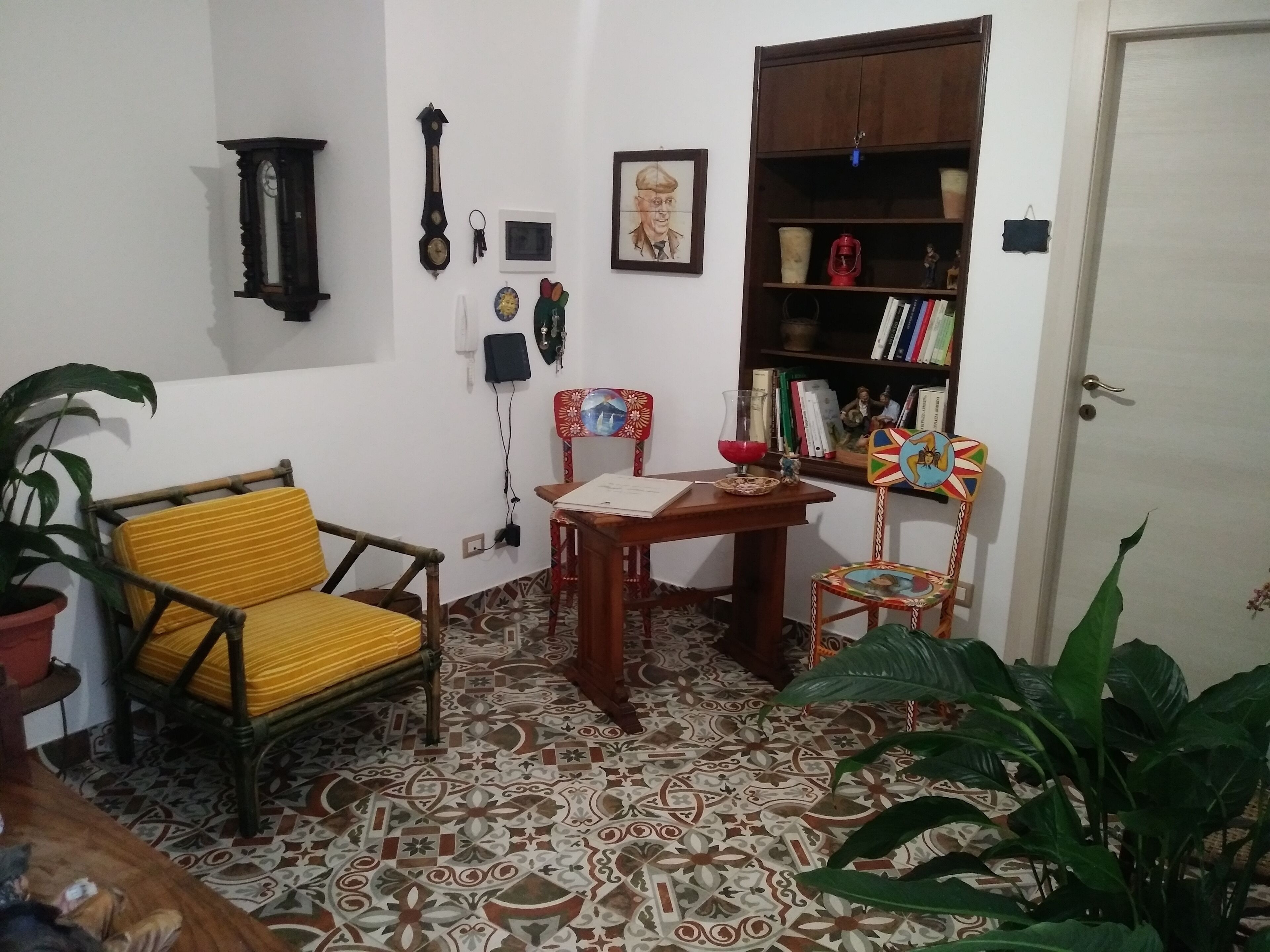 A room with a patterned tile floor, a yellow-striped chair, a wooden table with a book, a bookshelf, and a door.