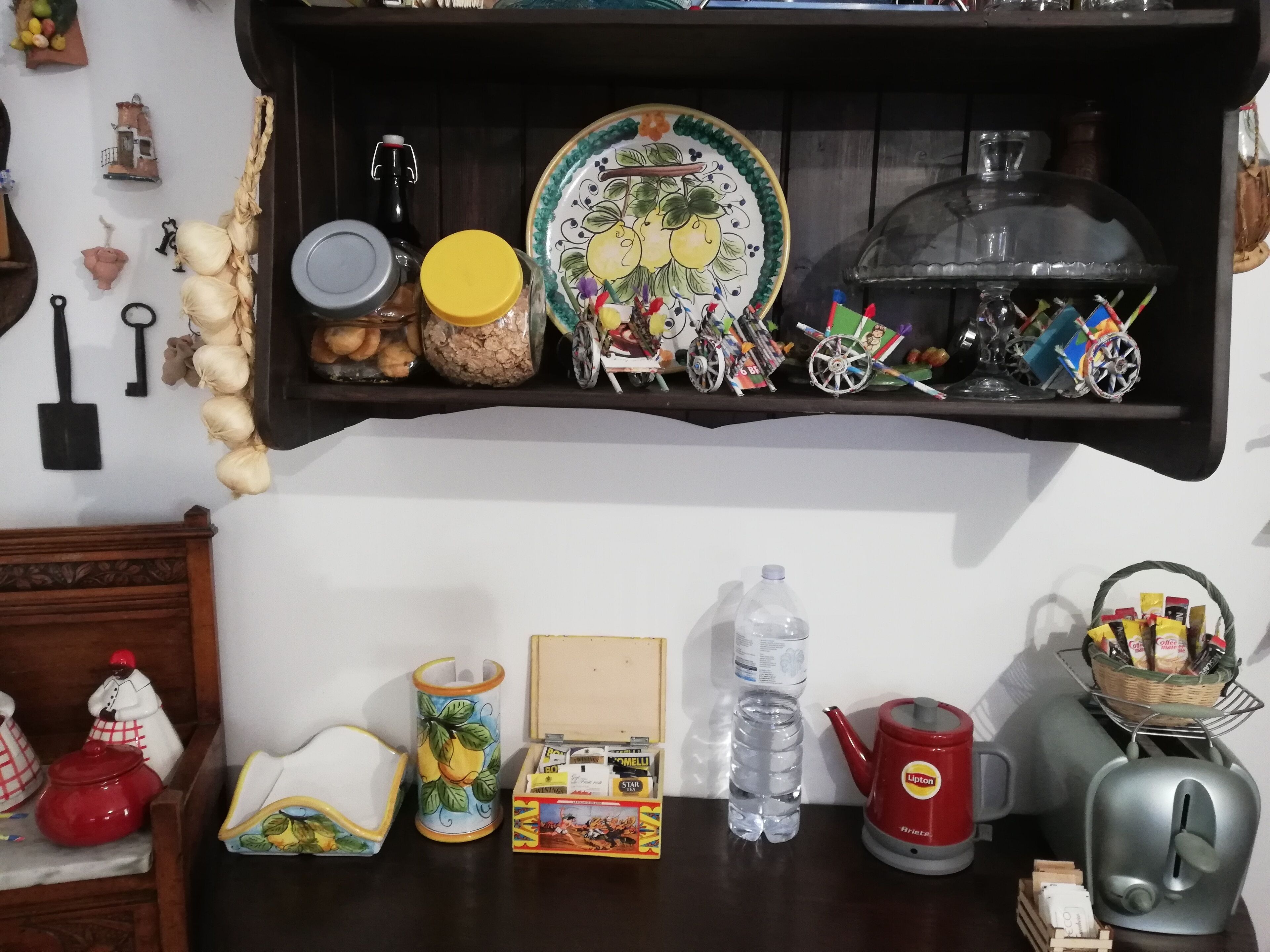 Un estante de cocina con varios objetos, incluyendo un plato con diseño de frutas, un frasco, una botella y una tetera.